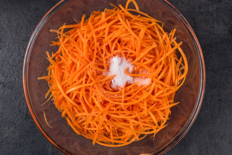 Grated carrots in a large bowl being mixed with salt, sugar, and vinegar to release juice.