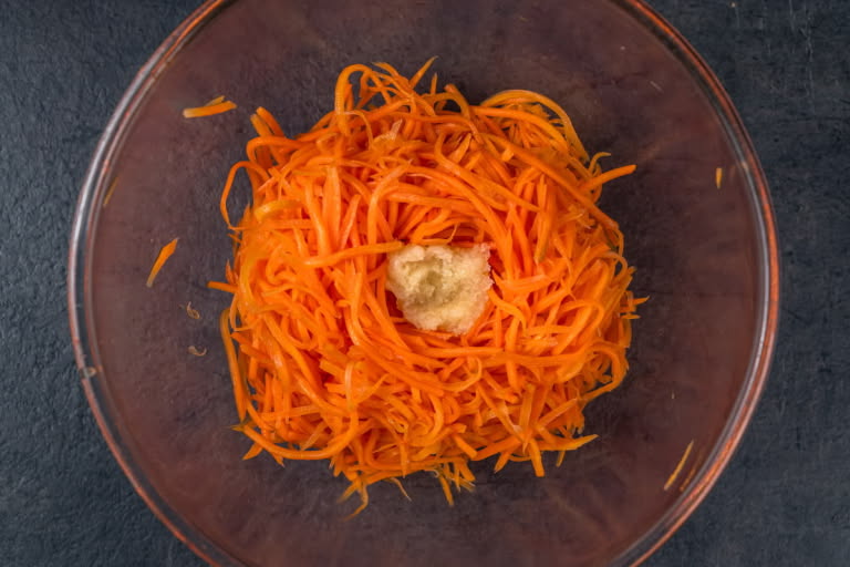 A mound of drained carrots in a bowl with a well in the center, and pressed garlic being added into the well.
