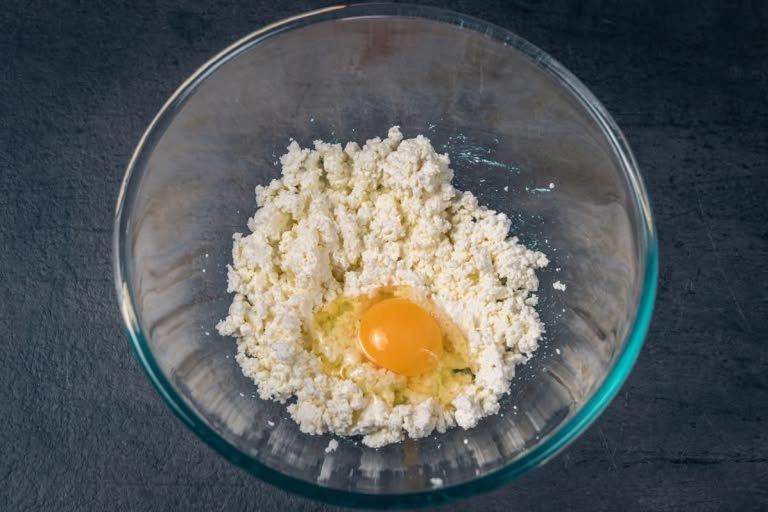 A medium egg cracked into the bowl with mashed farmer's cheese.